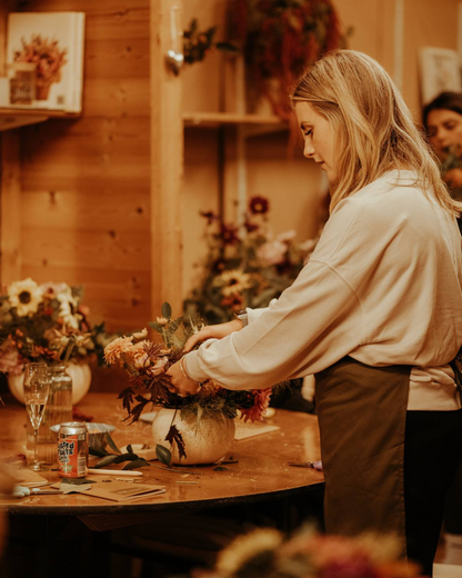 Woman arranging flowers in a vase at a wooden table with a warm, indoor setting.